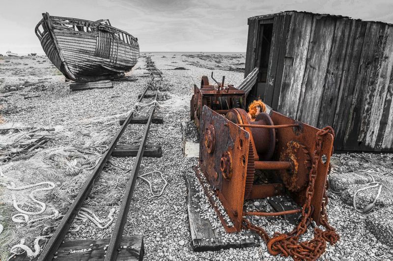 Old winch and fishing boats found at Dungeness which is a headland on the coast of Kent, England, formed largely of a shingle beach in the form of a cuspate foreland. It shelters a large area of low-lying land, Romney Marsh. Dungeness is also the name of the power station, of the hamlet within the location, and of an important ecological site at the same location.